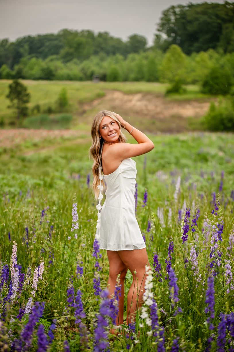 Senior girl wearing a White sundress contrasts with purple lupine flowers in a sunny meadow.