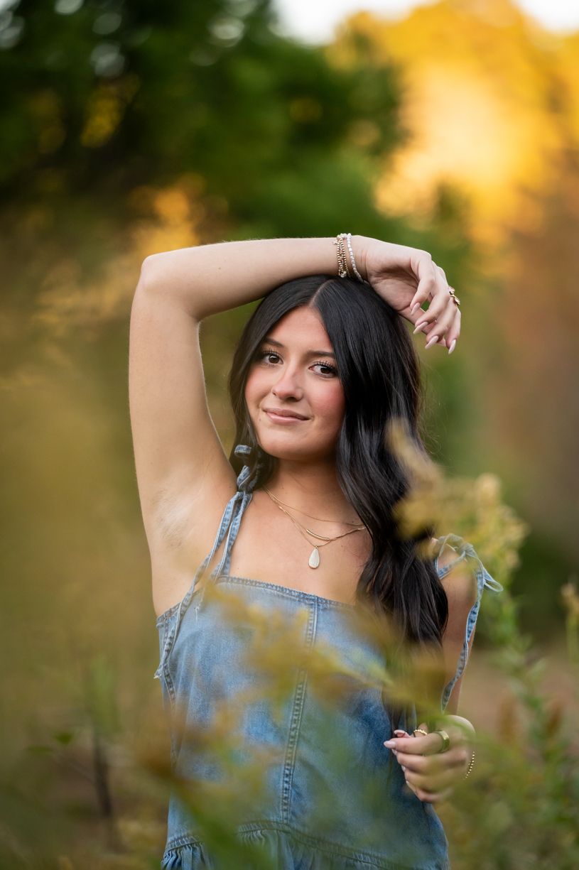 A senior girl Portrait series in denim overalls against golden sunset lighting in natural setting.