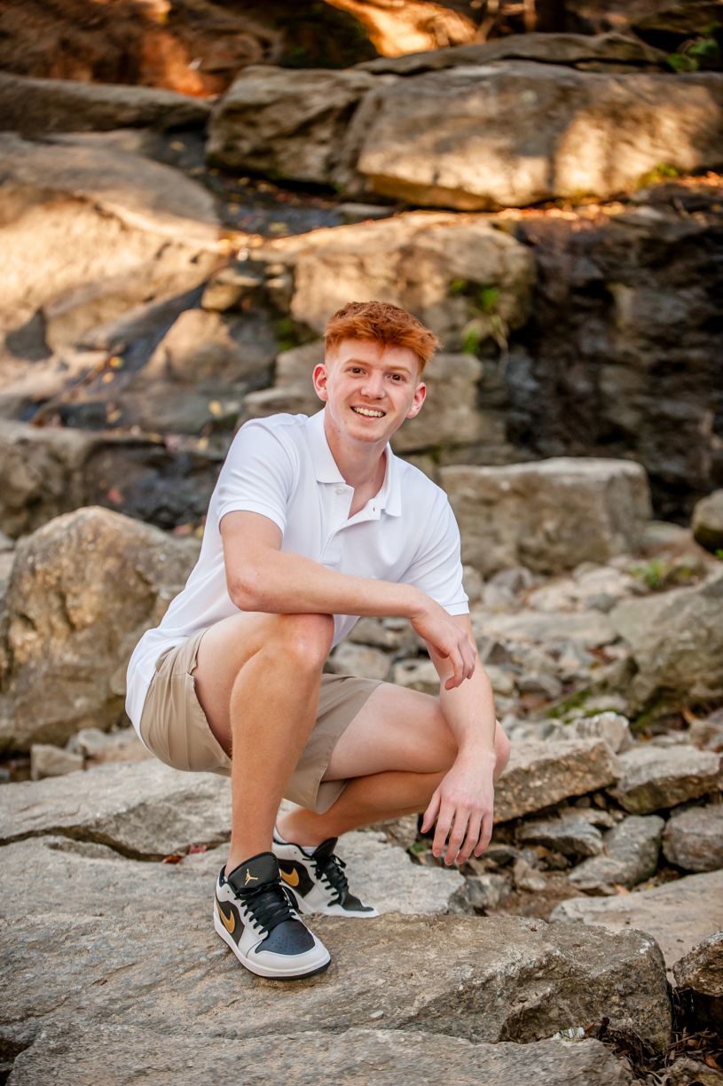 Senior guy poses casually on rocky terrain wearing white and khaki casual attire.