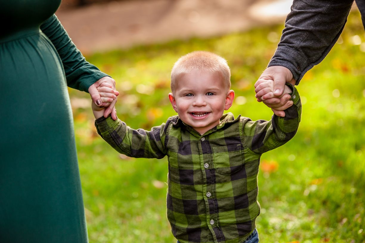 A small child in plaid holds hands with adults while smiling outdoors.