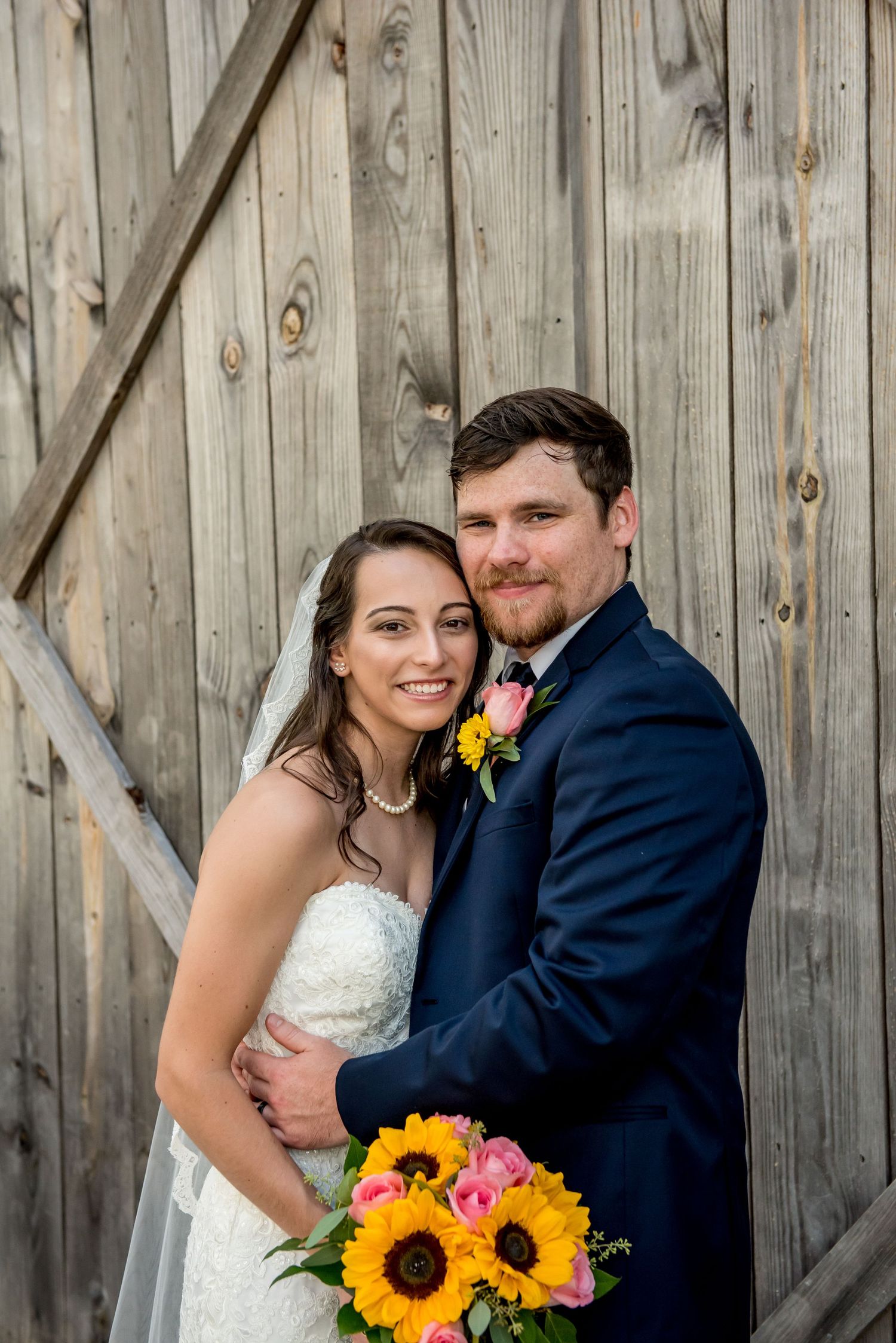 Bride and groom smiling together in front of rustic barn doors, bride holding vibrant sunflower and pink rose bouquet.
