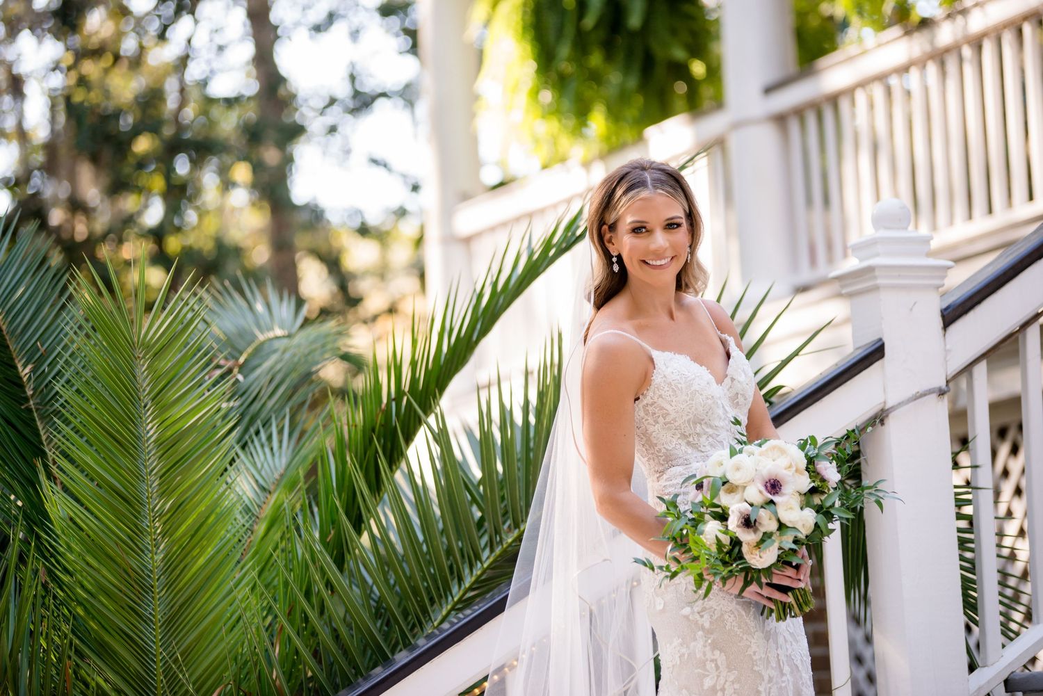 A beautiful bride in a lace wedding gown holds a floral bouquet while posing on elegant white stairs.