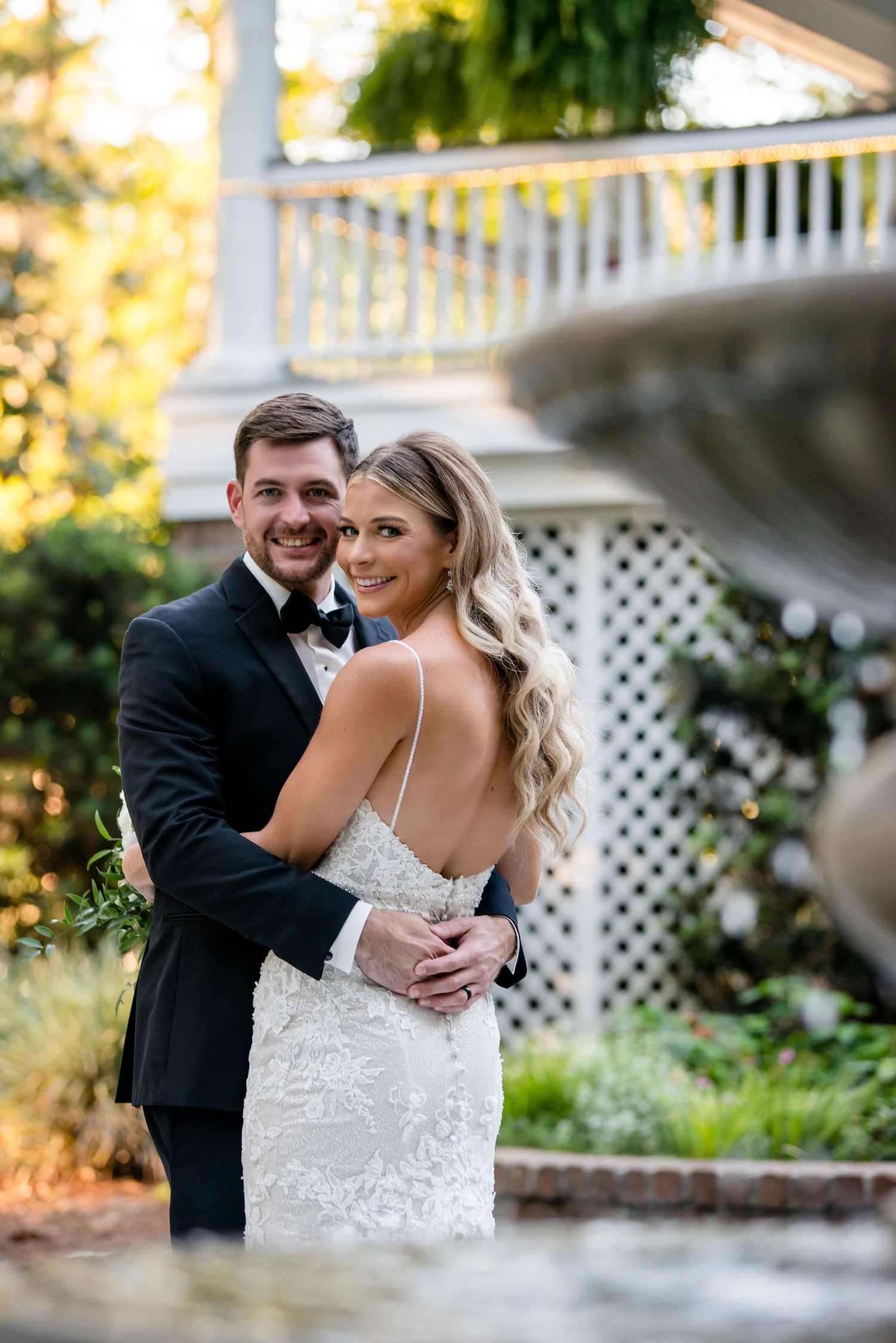 A smiling bride and groom embrace outdoors near a fountain, she in a backless lace gown, he in a black tuxedo.