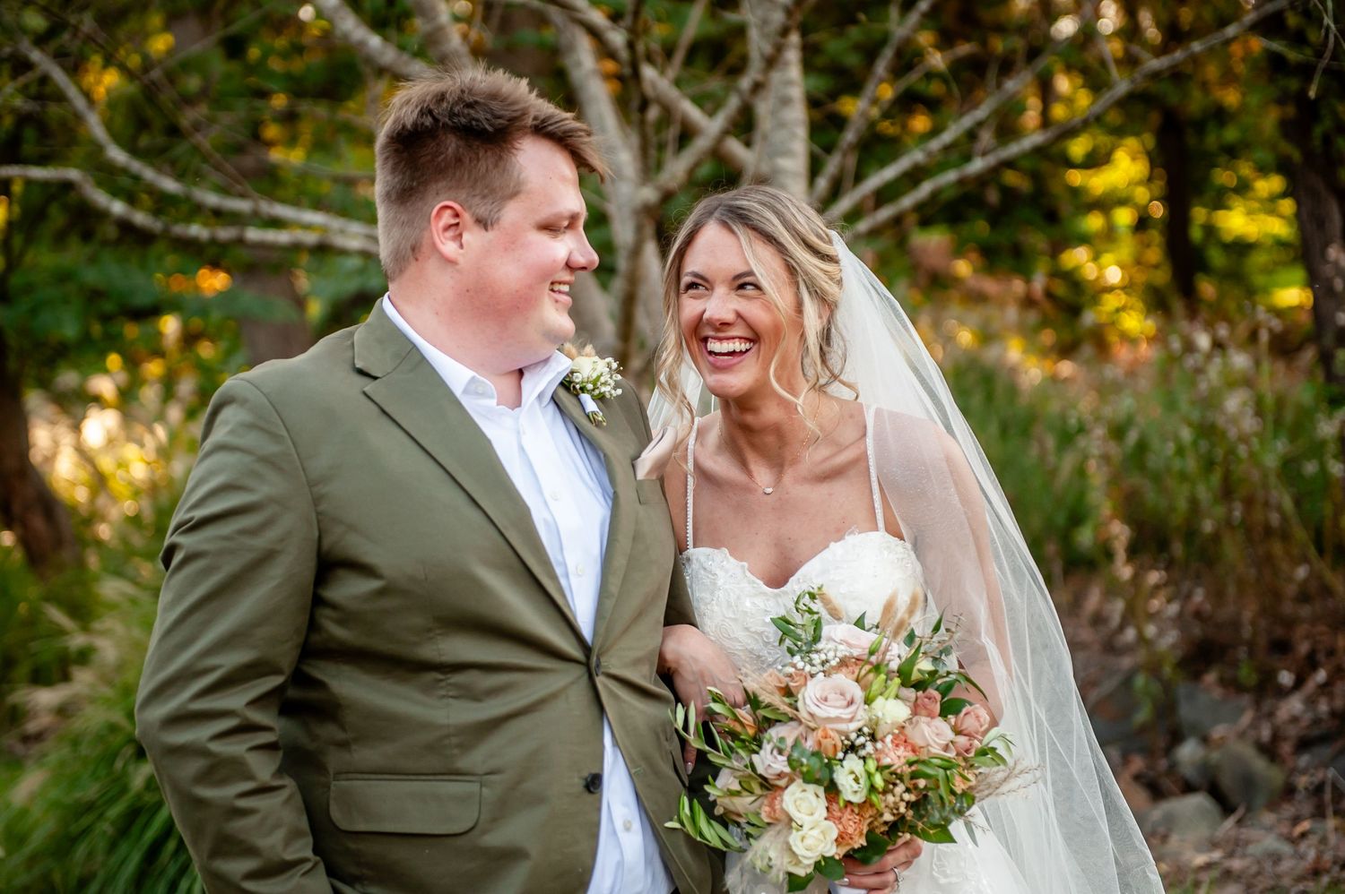 Smiling bride in white gown holding pink bouquet stands with groom in olive suit outdoors at wedding.