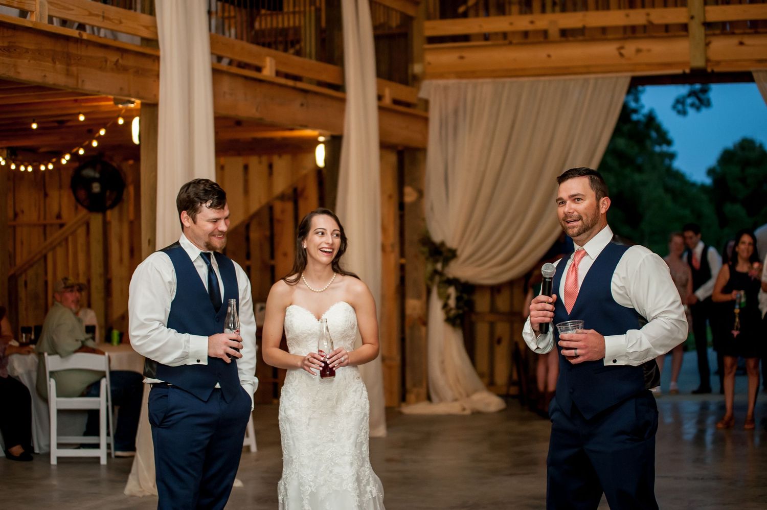Bride in white gown stands with two groomsmen in navy suits during a rustic barn wedding reception.
