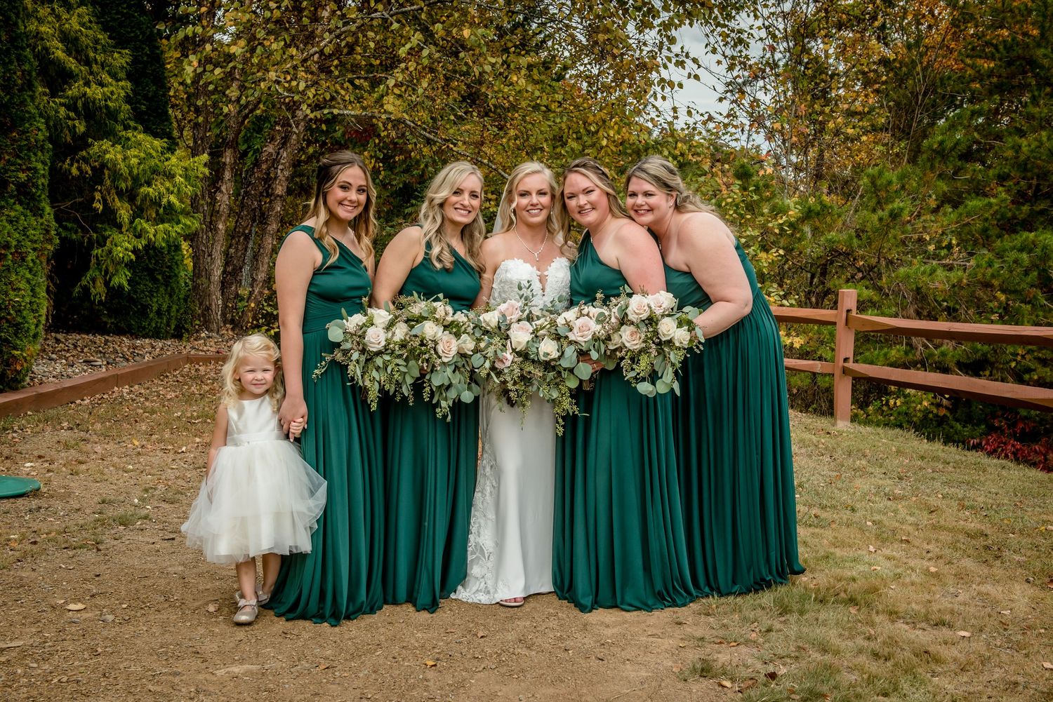Bride in white gown with four bridesmaids in emerald green dresses holding bouquets, posing outdoors with a flower girl.