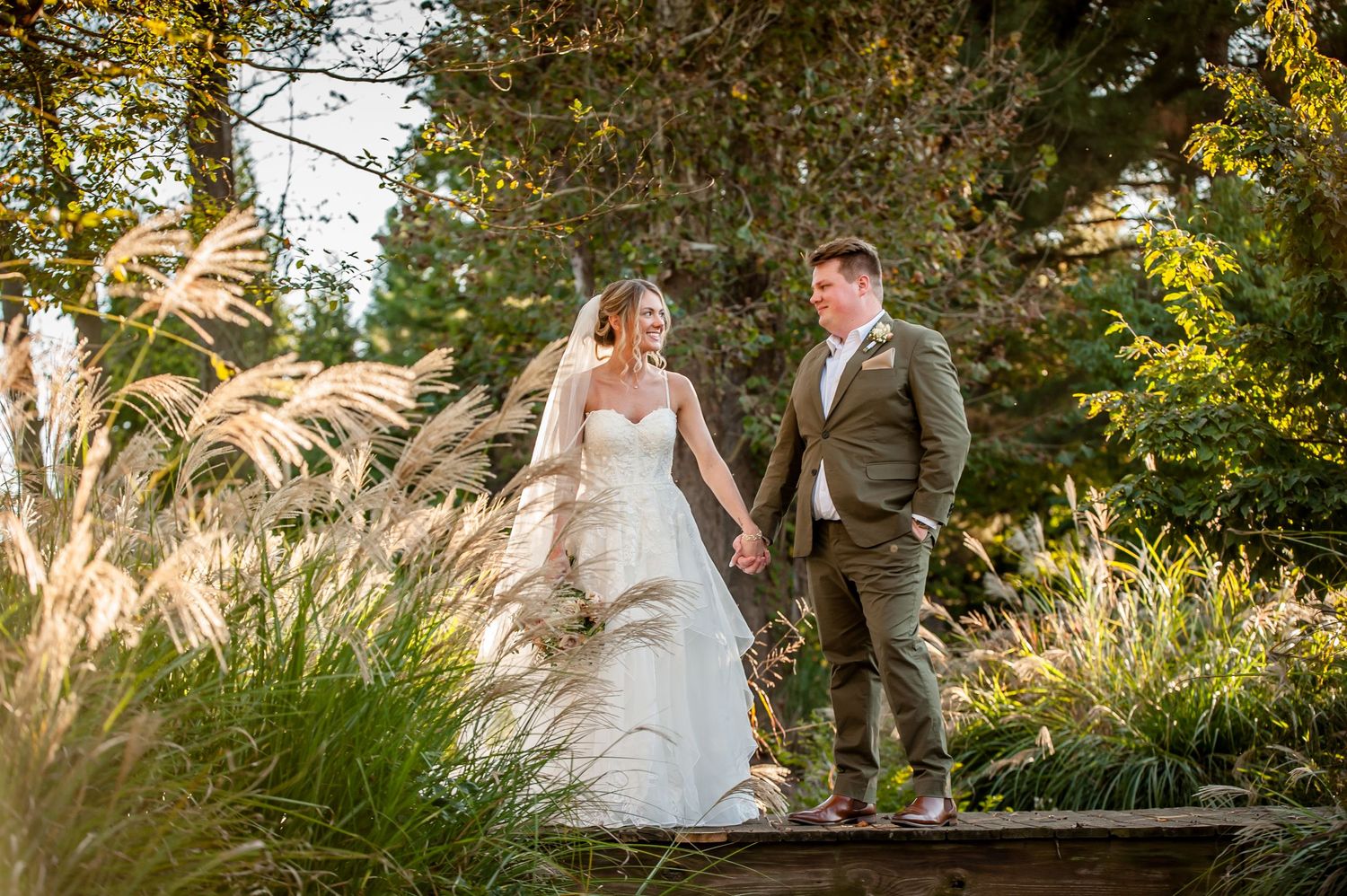 Bride and groom hold hands walking through lush garden greenery in golden sunlight on their wedding day.