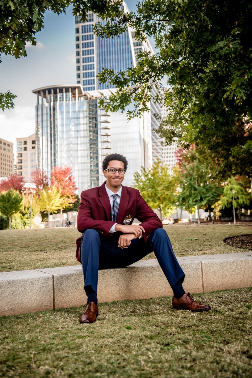 A senior boy wearing in burgundy blazer sitting on stone wall with city skyline background.