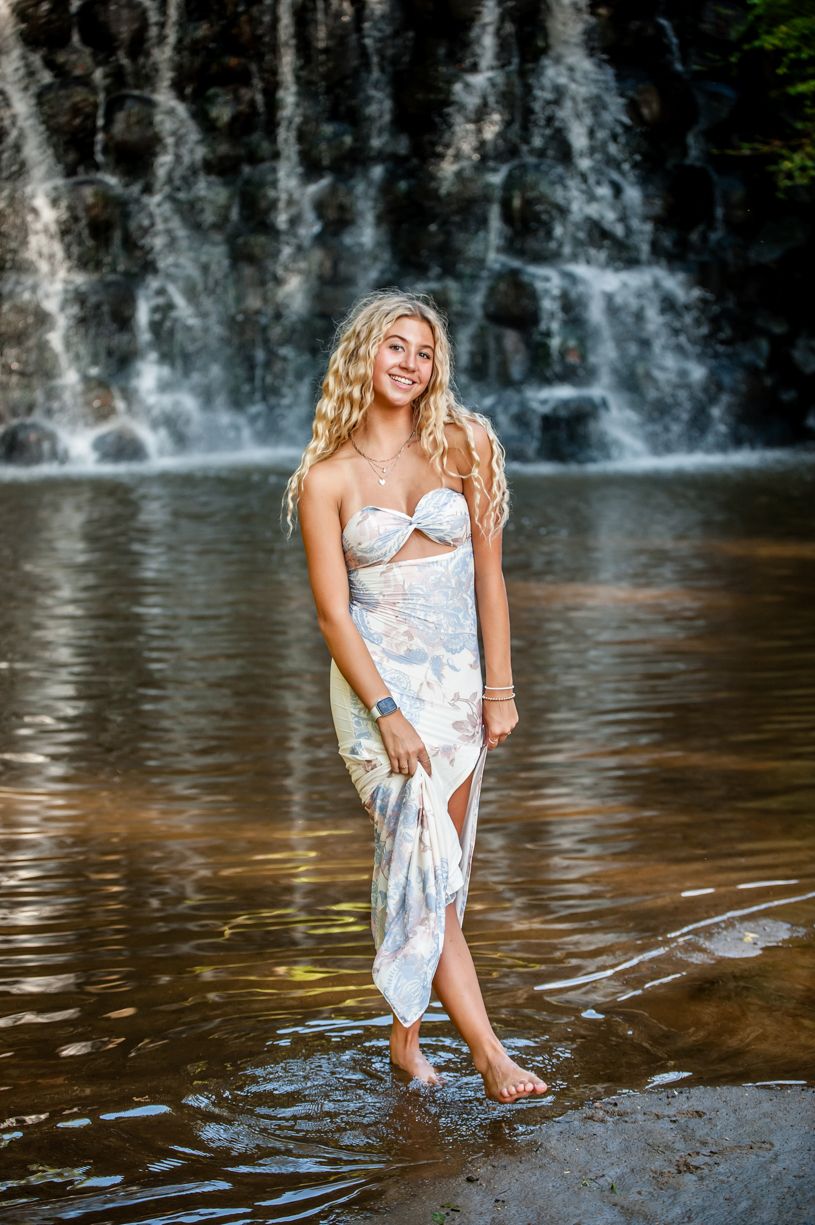 Senior girl posing by a waterfall wearing a light-colored summer dress.