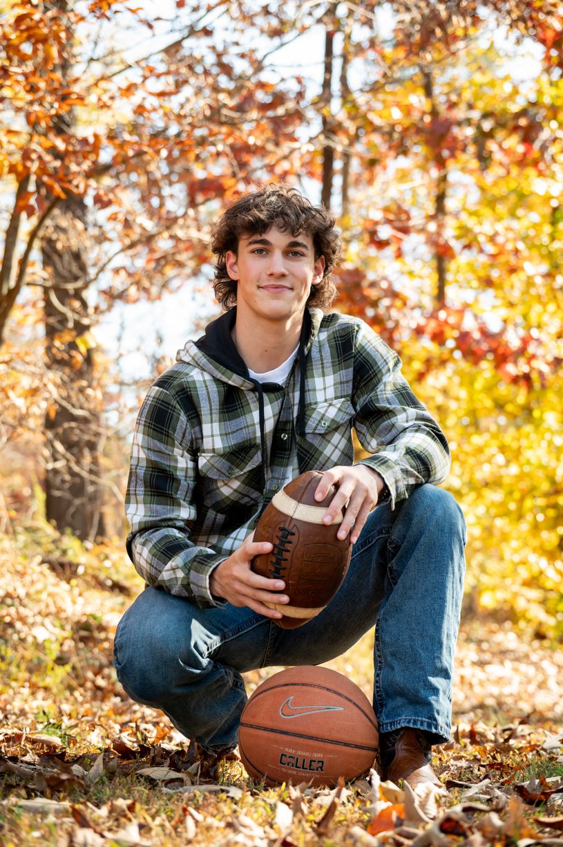 High school senior poses with basketball and football in autumn forest setting wearing plaid shirt and jeans.