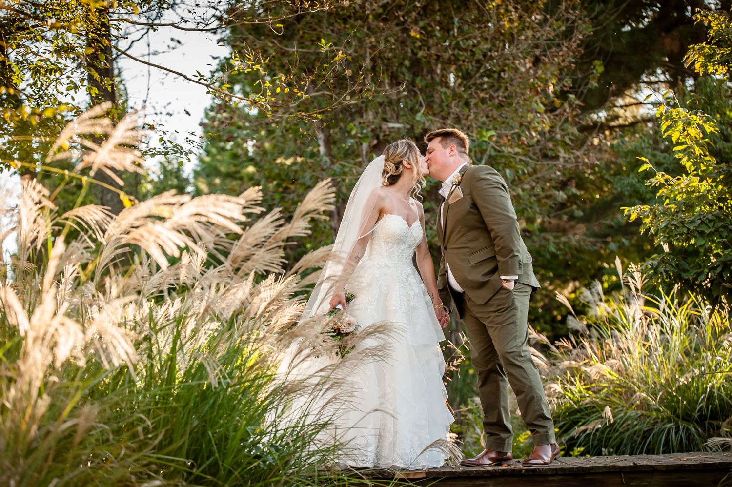 Bride and groom share a romantic kiss surrounded by tall ornamental grasses and lush greenery on their wedding day.