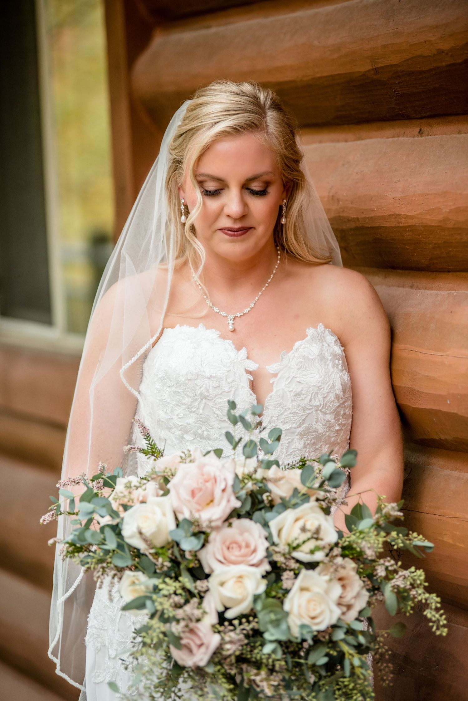 Bride in strapless lace wedding gown and veil holds blush rose bouquet against rustic log wall.