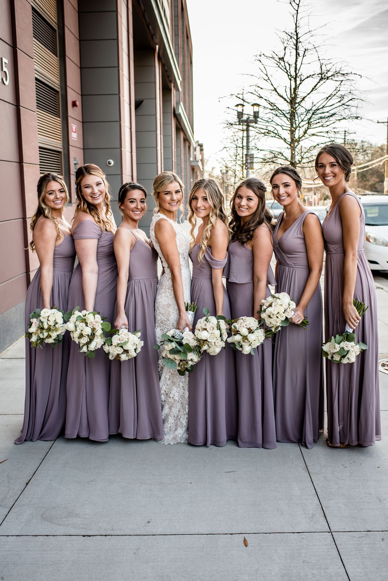 Bride and six bridesmaids in dusty purple gowns holding white floral bouquets pose outdoors on a sidewalk.