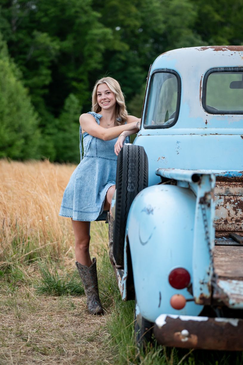 A vintage light blue pickup truck parked in a rustic field setting with a senior girl leaning against it.