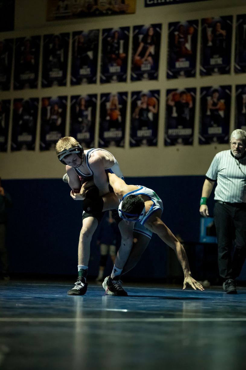 Wrestling match in progress on a gymnasium floor with referees observing.