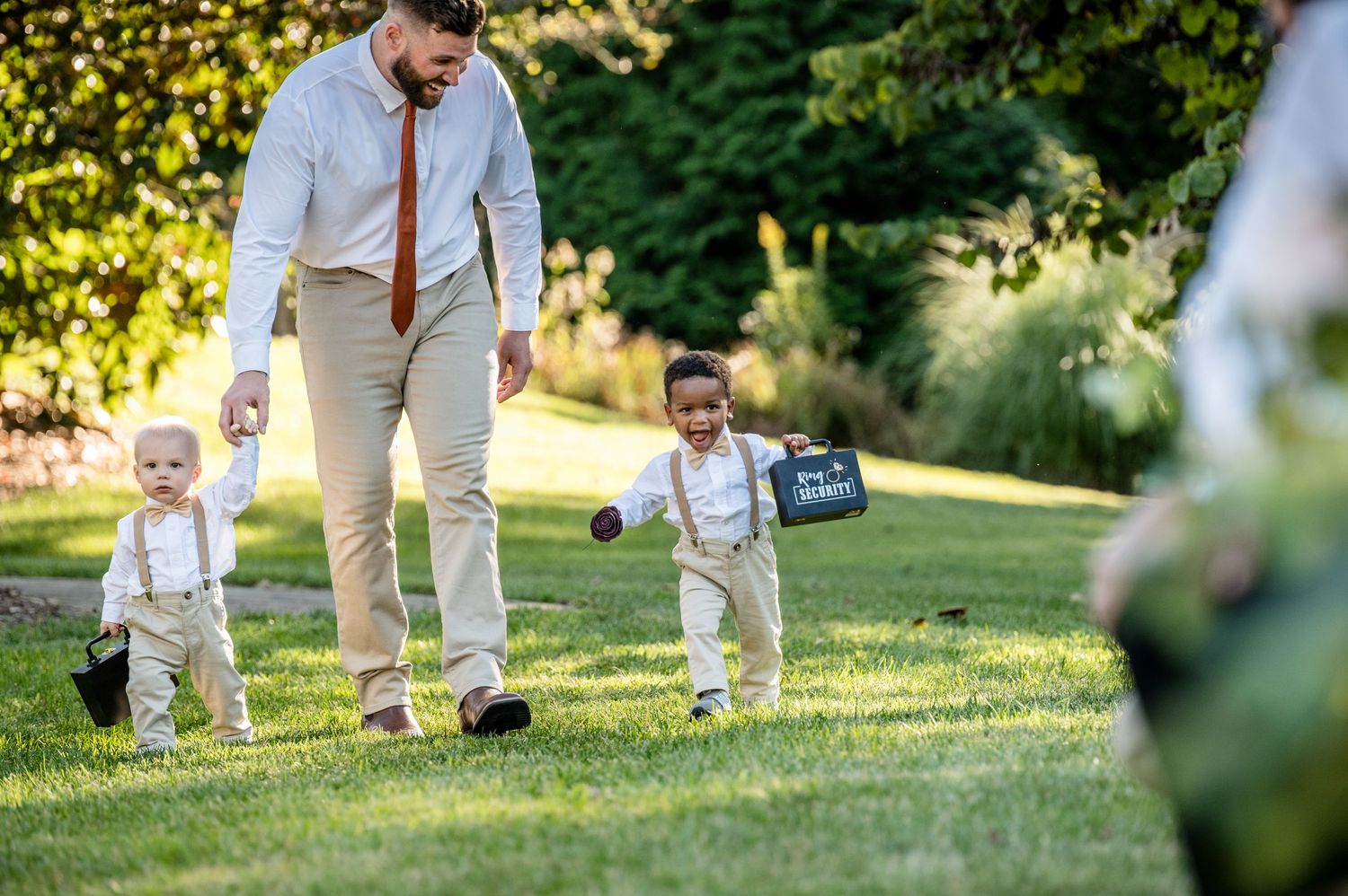 Man in white shirt walks with two toddlers in suspenders on green grass at an outdoor wedding.