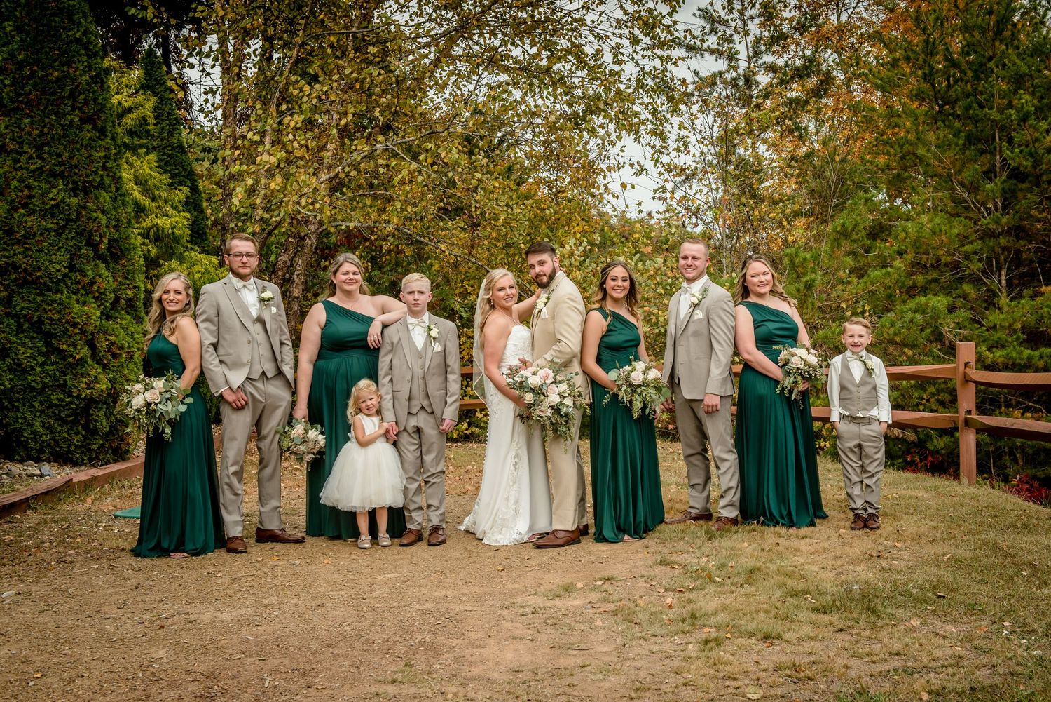 Wedding party in green dresses and tan suits posing outdoors near a wooden fence surrounded by lush trees.