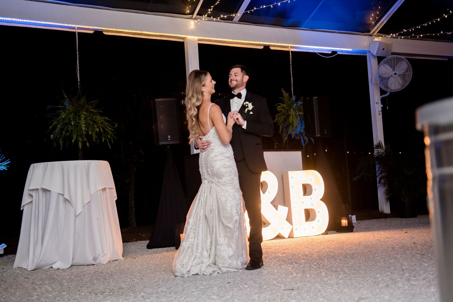 Bride and groom share first dance outdoors at night under twinkling lights with illuminated letter marquee signs.