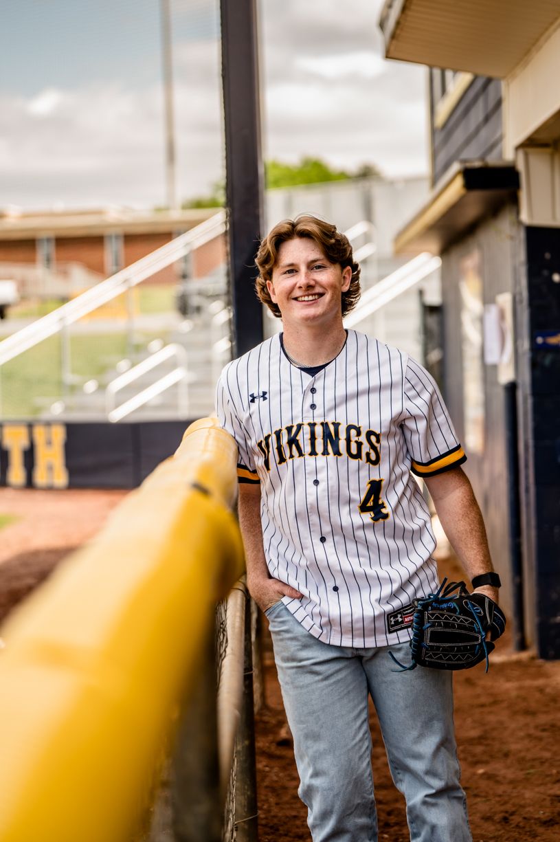 Senior Baseball player in Vikings pinstripe uniform poses on field near dugout.