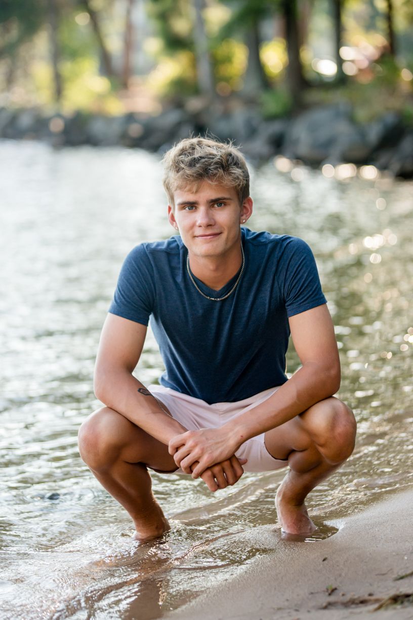 A senior guy in a black t-shirt crouching at the water's edge in a series of artistic poses.