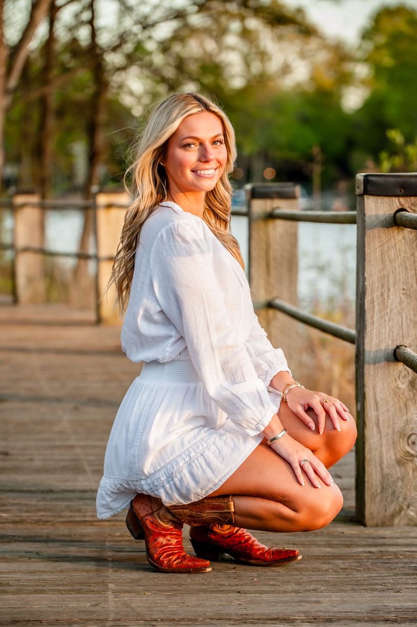 Senior girl in white dress poses on wooden dock wearing brown cowboy boots at sunset.