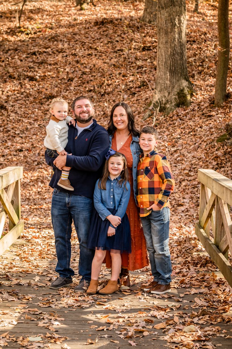 Autumn family portrait of five people standing together on wooden bridge surrounded by fall foliage.