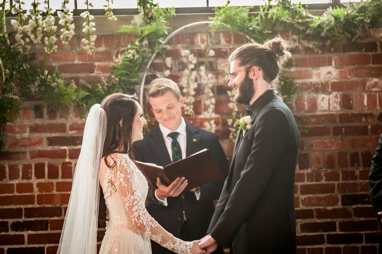 Bride and groom exchange vows before officiant at rustic brick wall wedding ceremony with circular floral arch backdrop.