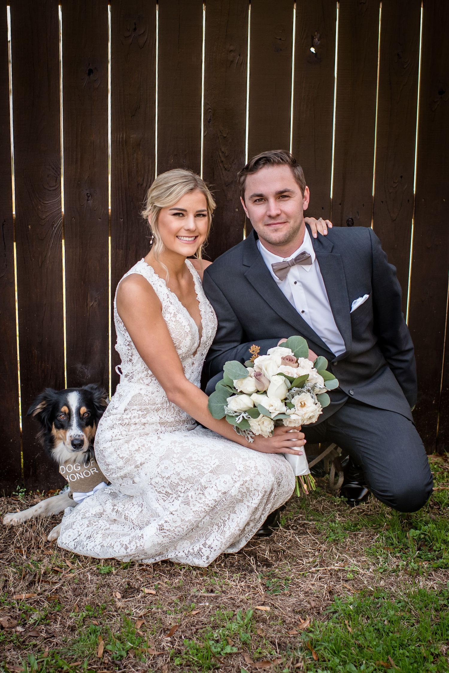 Bride and groom posing with their dog in front of a wooden fence, bride holding white floral bouquet.