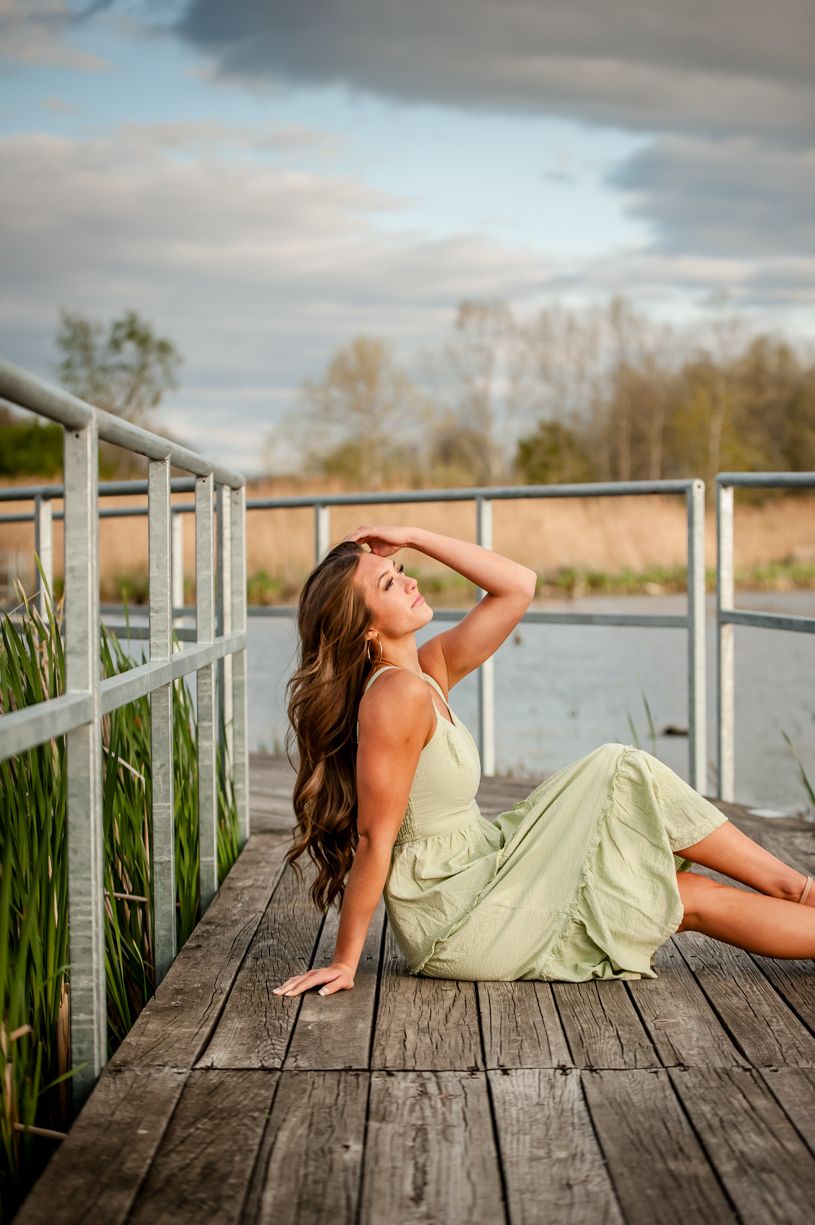 Senior girl in sage green dress poses dramatically on wooden dock during golden hour.