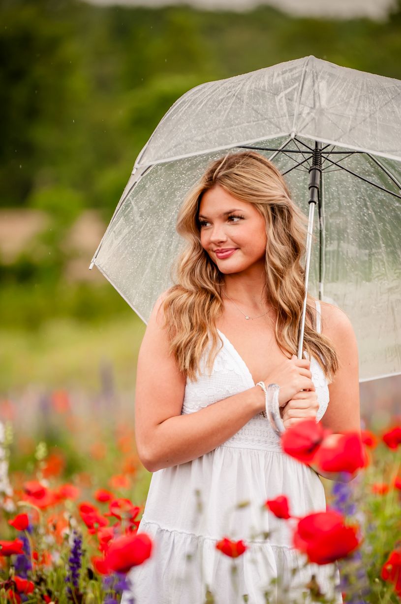 Senior girl Portrait with clear umbrella among red poppies and wildflowers on a cloudy day.
