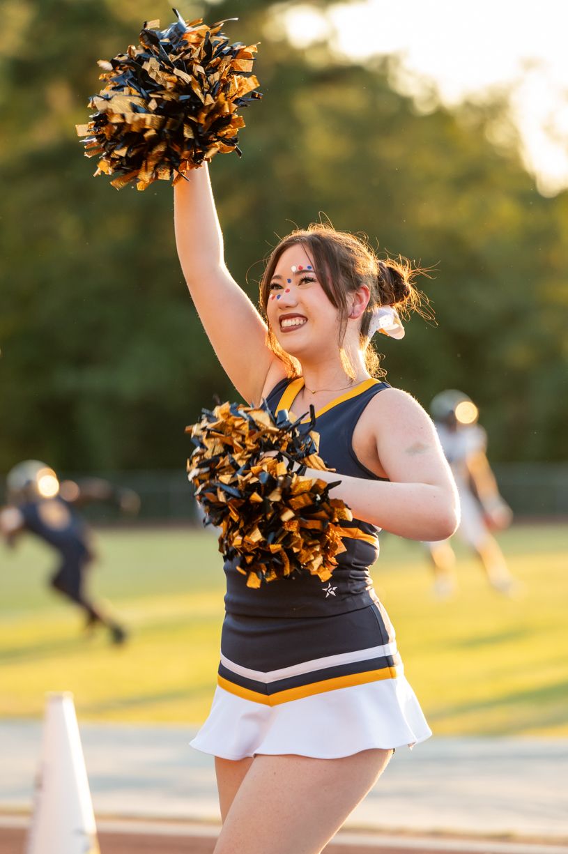 Cheerleader in navy and gold uniform raising pom pom during outdoor performance.