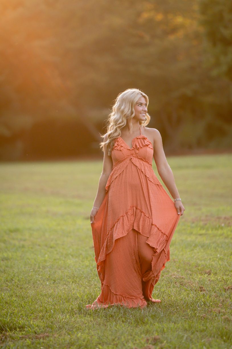 A senior girl wearing a flowing coral dress captured in golden hour on grass.