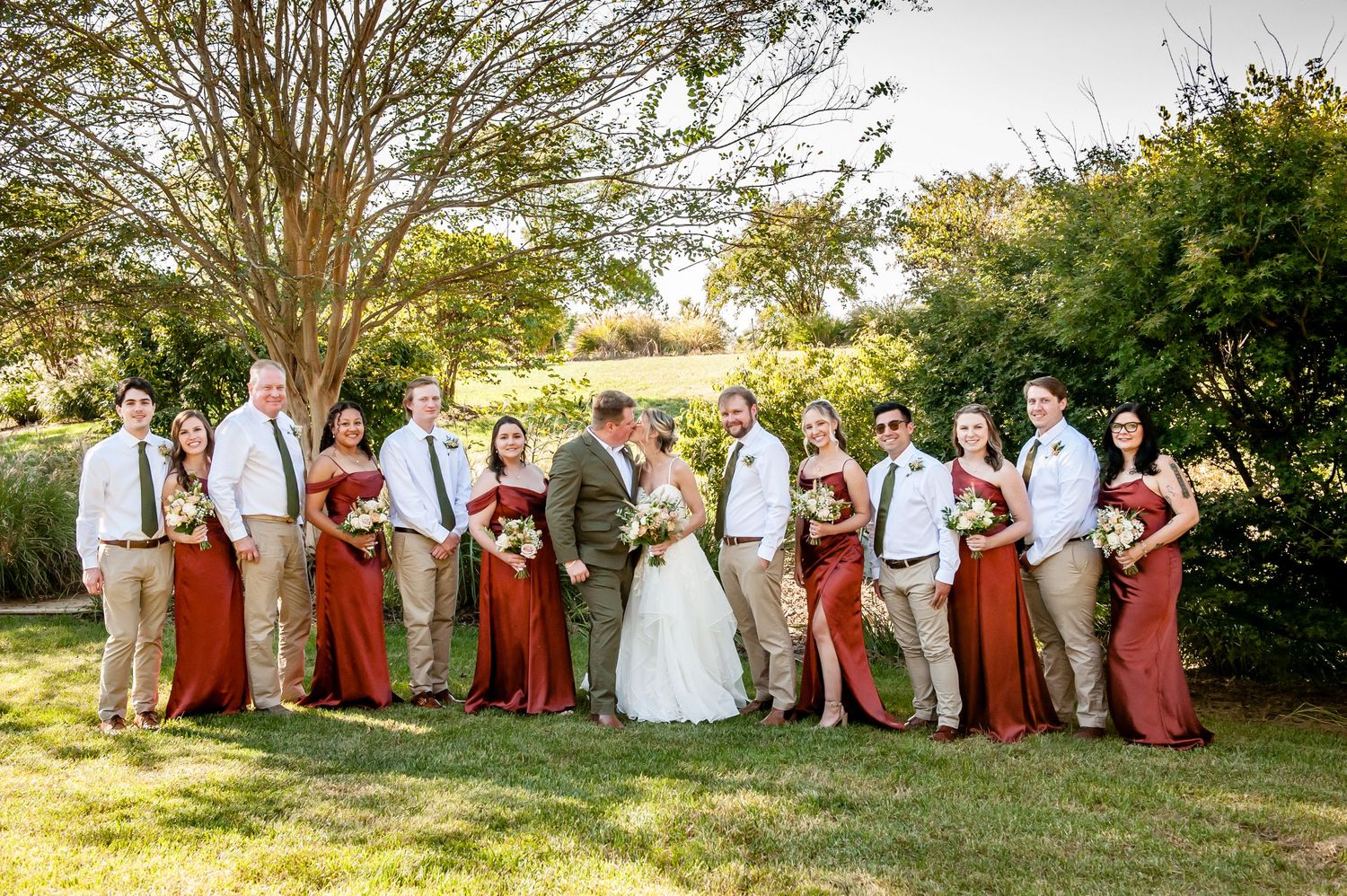 Wedding party poses outdoors, bridesmaids in red dresses, groomsmen in tan suits, bride and groom centered on lush green lawn.