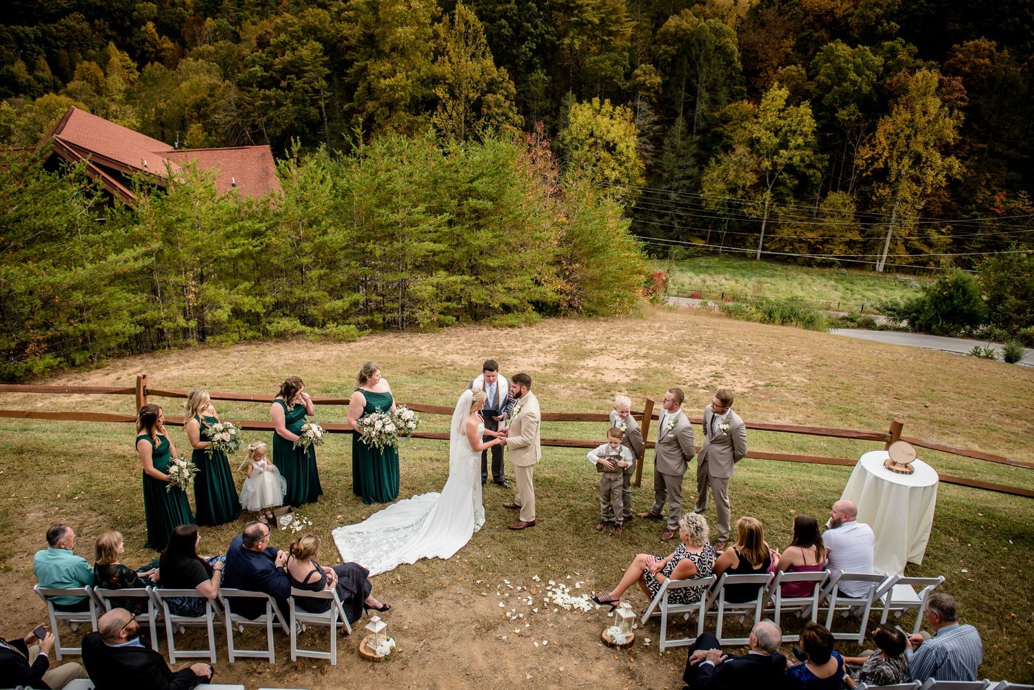 Outdoor wedding ceremony on a hillside with bridal party in green dresses, surrounded by autumn trees and rustic barn backdrop.