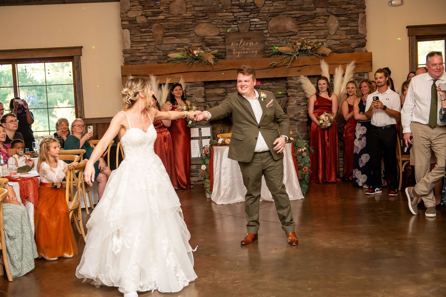 Bride and groom share their first dance at rustic wedding reception with guests applauding in background.