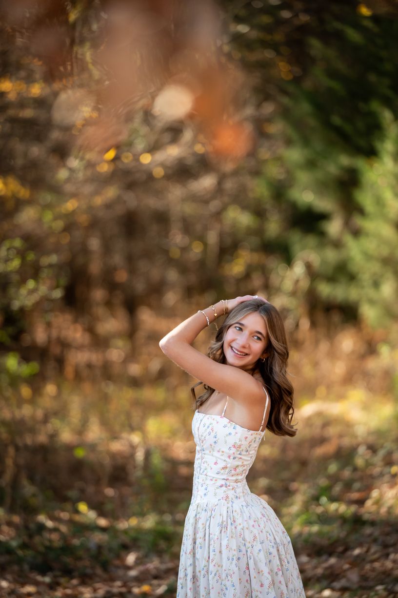 A senior girl in white floral dress twirls playfully among fall foliage.