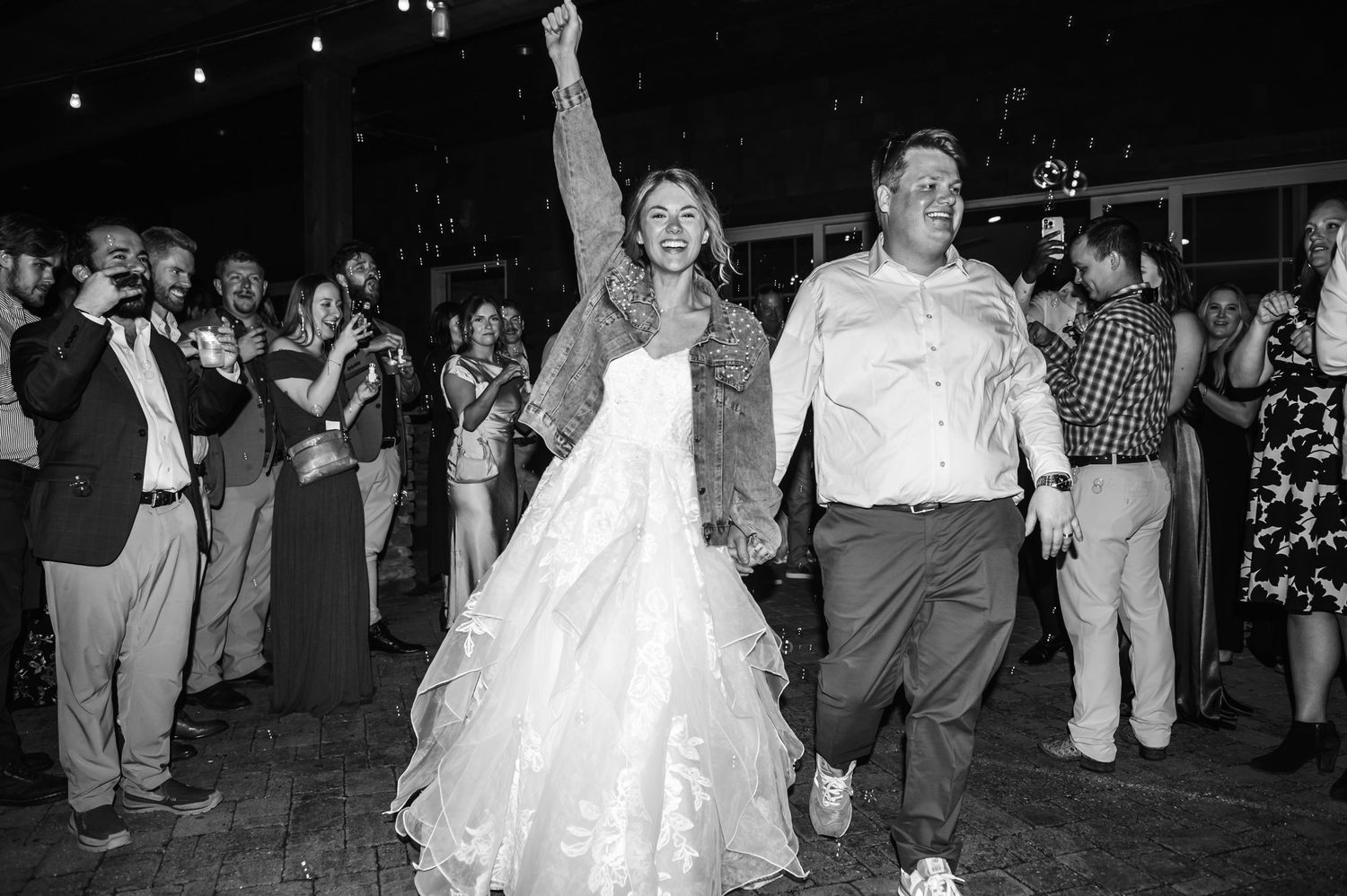 Bride in wedding gown and denim jacket celebrates with groom on dance floor surrounded by cheering wedding guests.