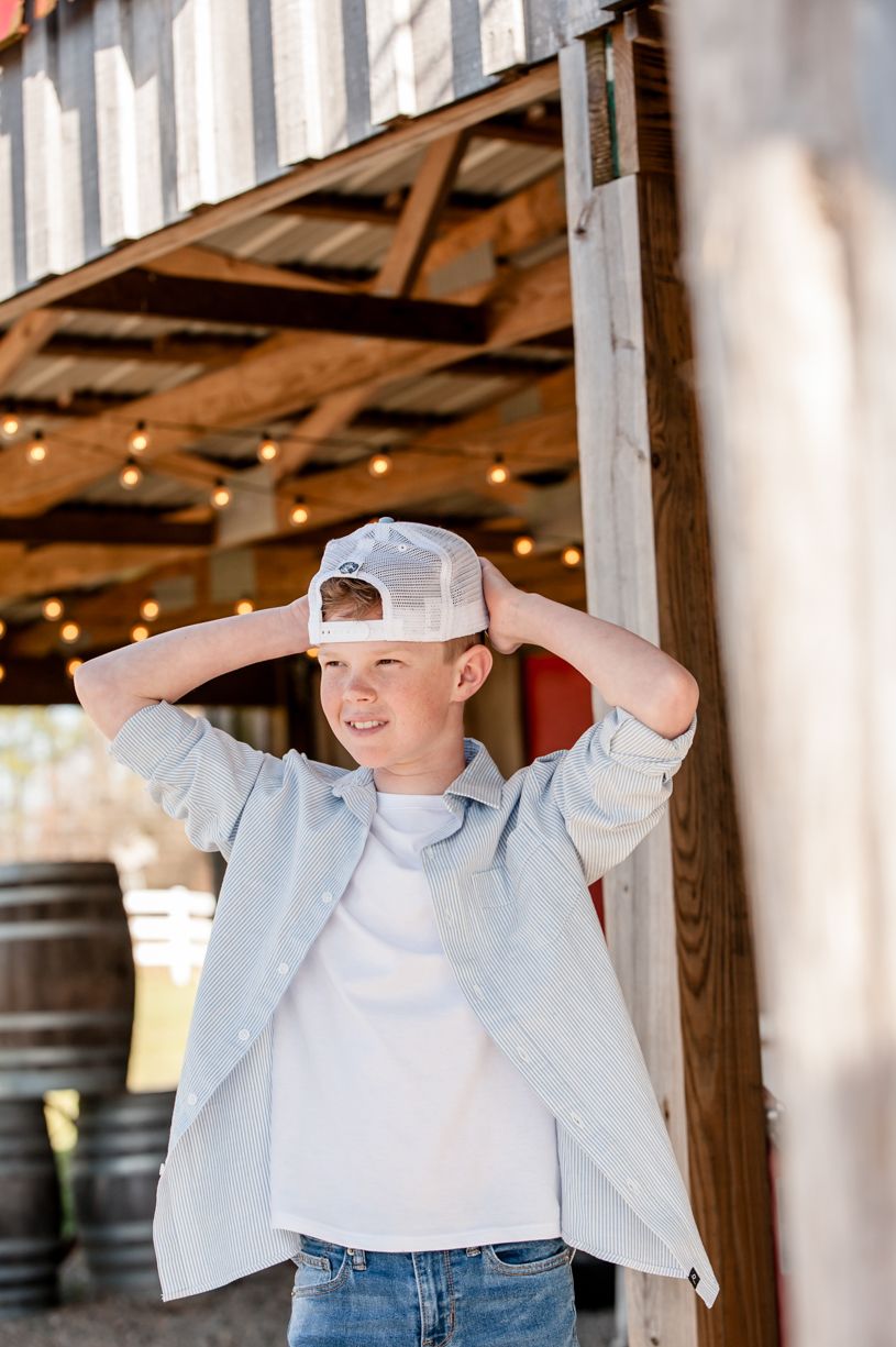 Young person in casual attire adjusting white baseball cap under rustic wooden structure.
