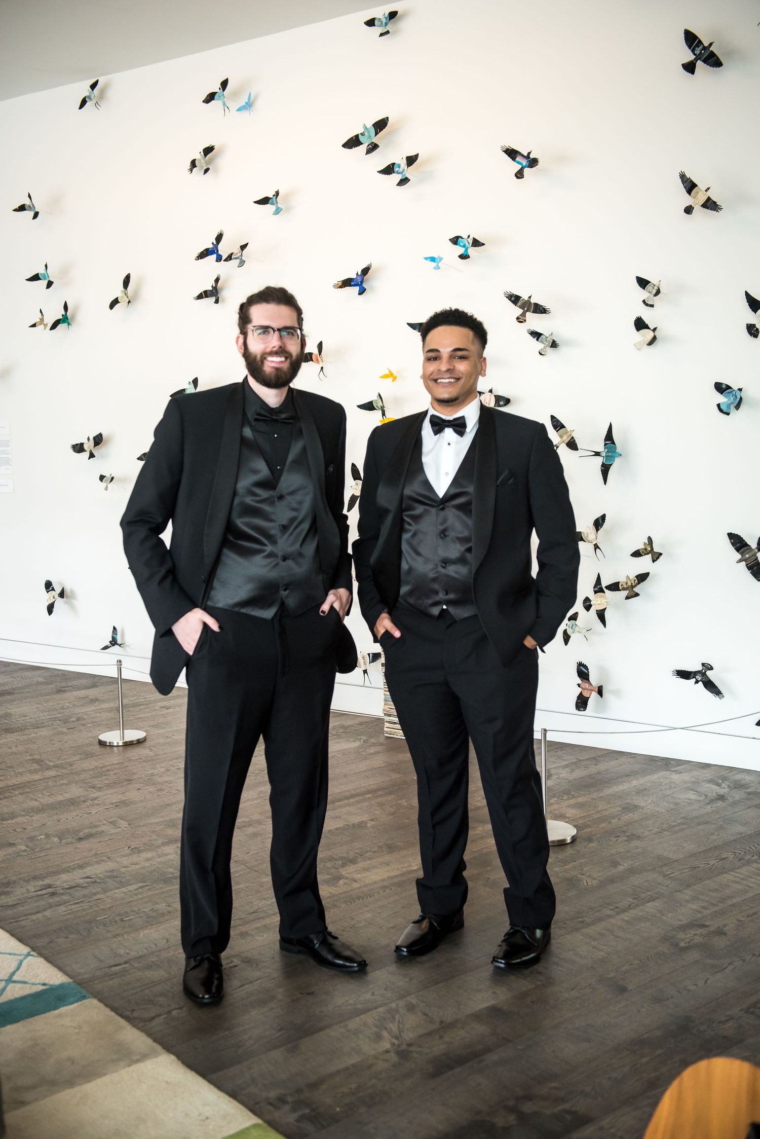 Two men in black tuxedos pose in front of a white wall adorned with colorful butterfly sculptures at a formal event.