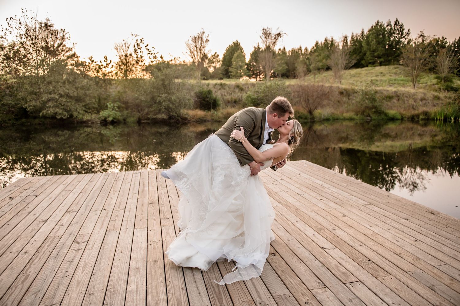 Bride and groom share romantic dip kiss on wooden dock overlooking serene pond at golden hour wedding.