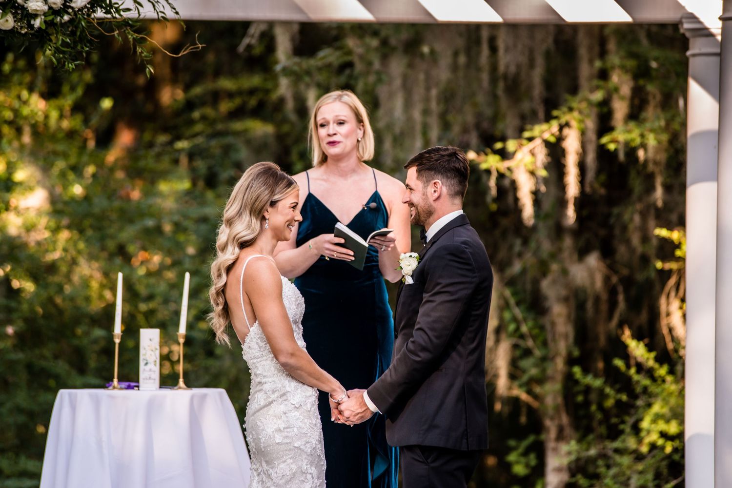 Bride and groom hold hands during outdoor wedding ceremony while officiant in blue velvet dress reads vows under pergola.