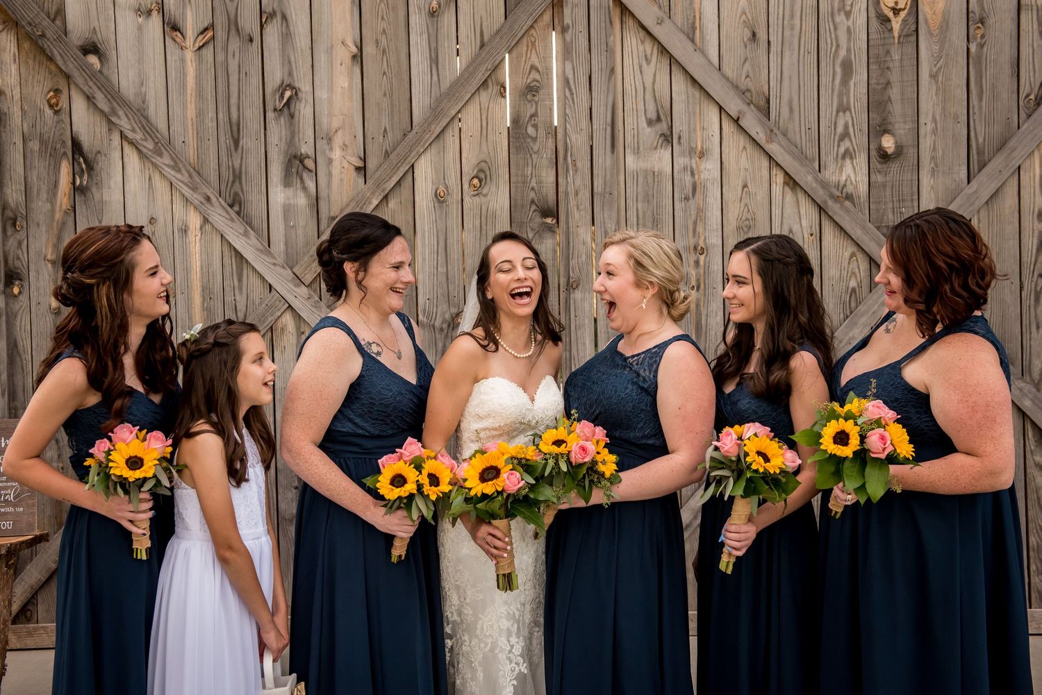 Bride laughing with bridesmaids in navy dresses holding sunflower bouquets in front of rustic barn doors.