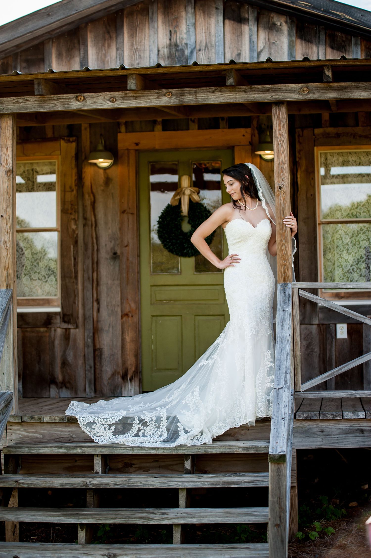 Bride in strapless lace mermaid gown with cathedral veil poses on rustic wooden porch with green door.