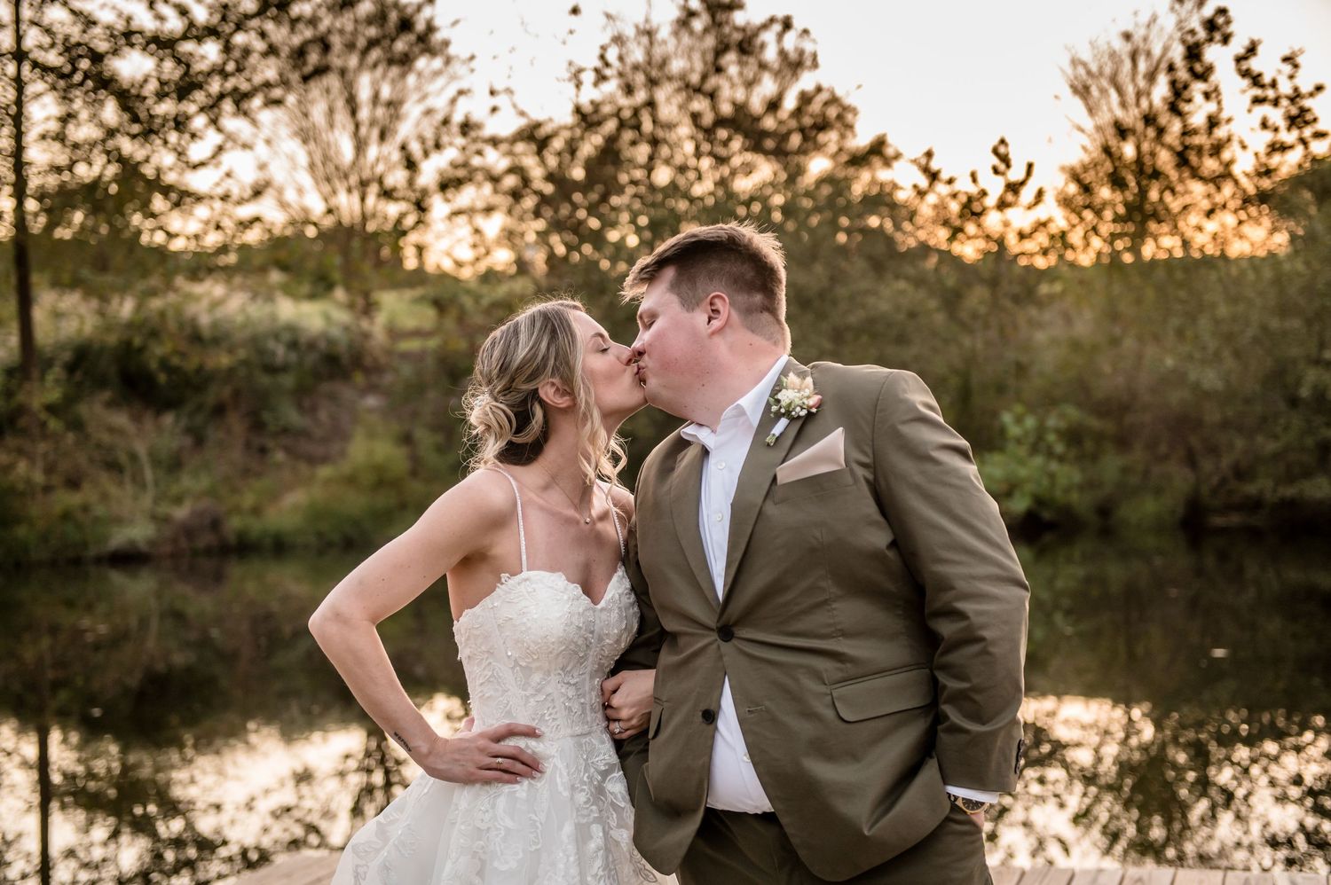 Bride and groom share a romantic kiss at golden hour beside a serene pond, groom in tan suit, bride in lace gown.