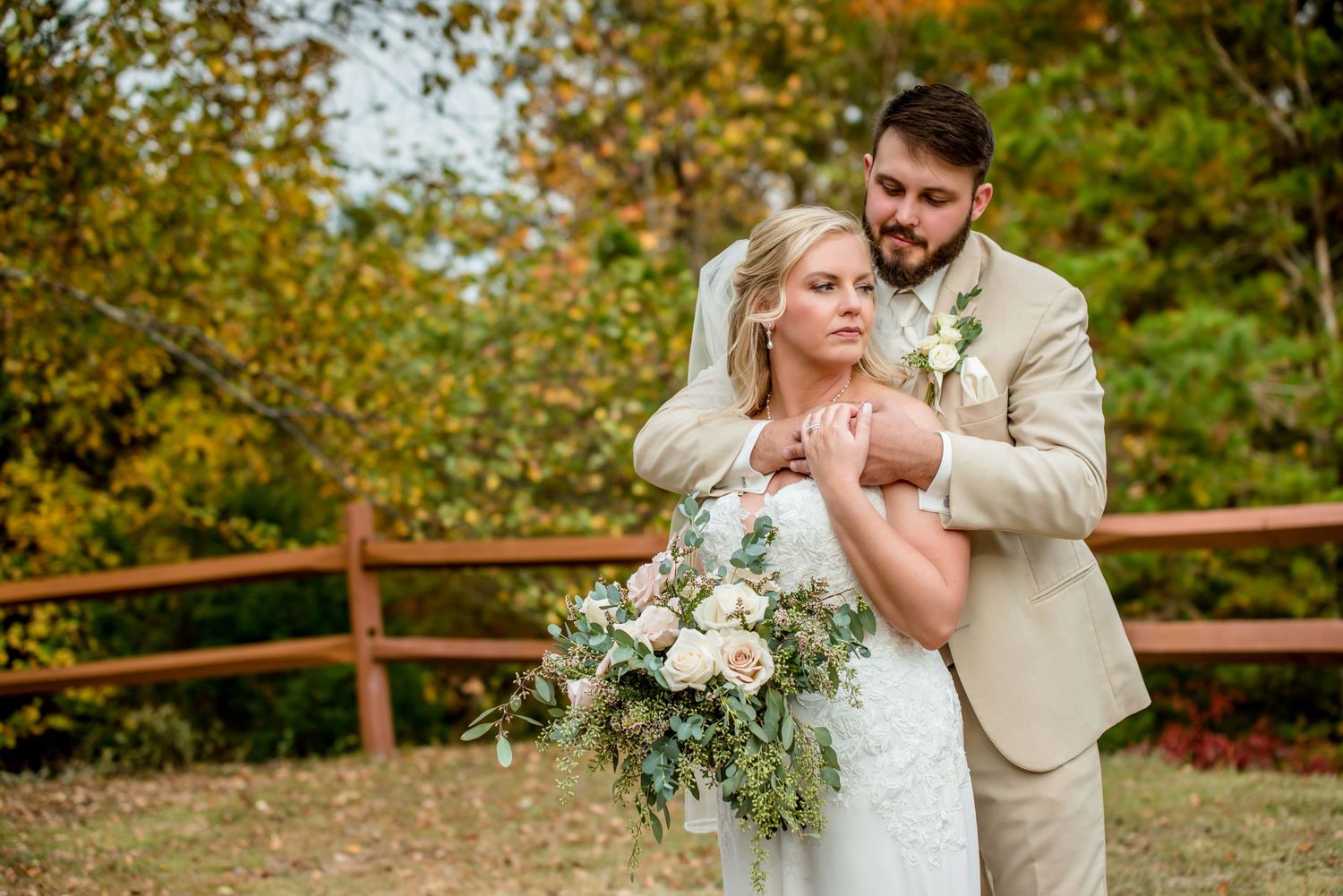 Bride and groom embrace outdoors in fall foliage, she holds a white floral bouquet wearing a white gown, he wears a tan suit.