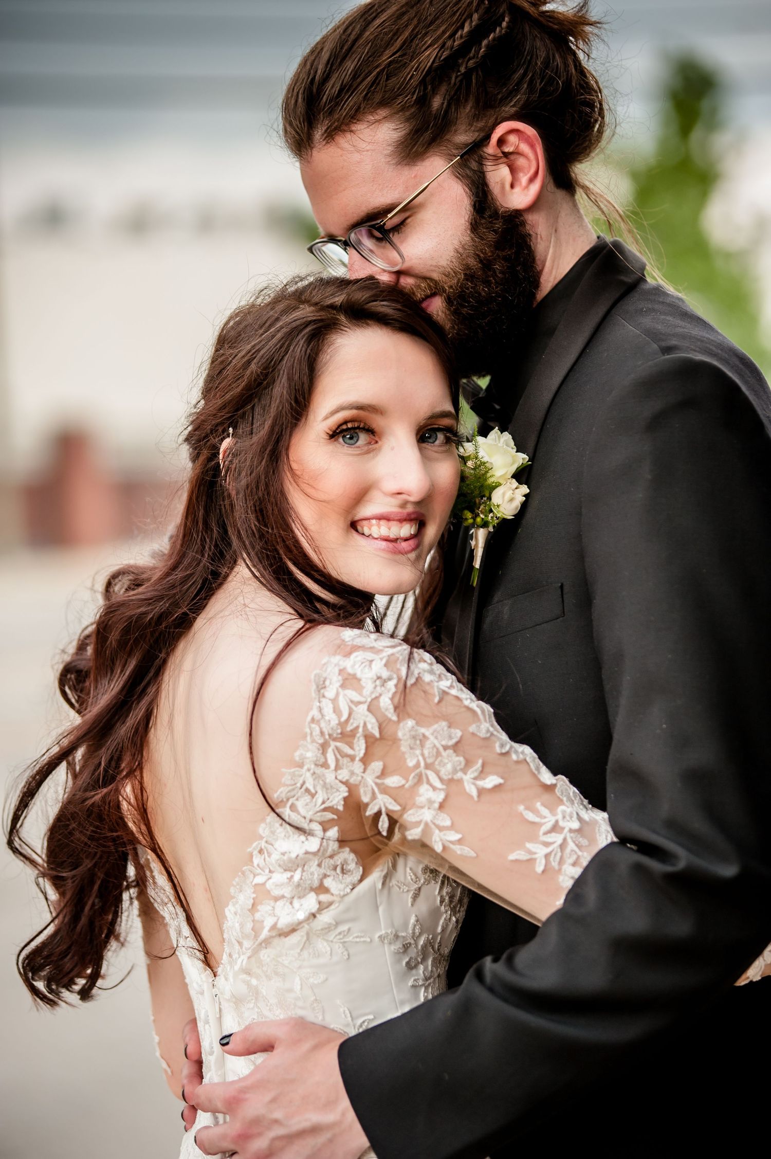 Bride in lace gown smiles as bearded groom in black suit kisses her head during romantic wedding portrait.