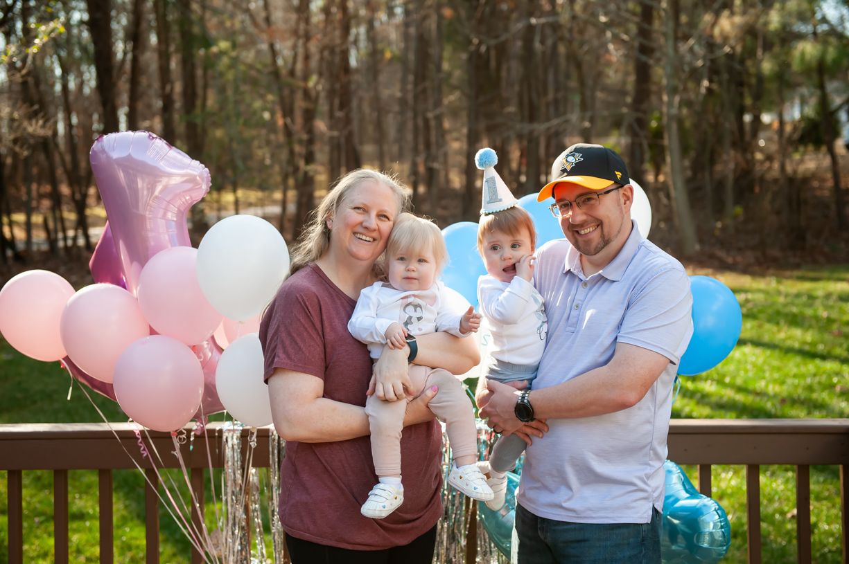 A family celebration on a wooden deck with pink and blue balloons in a forest setting.