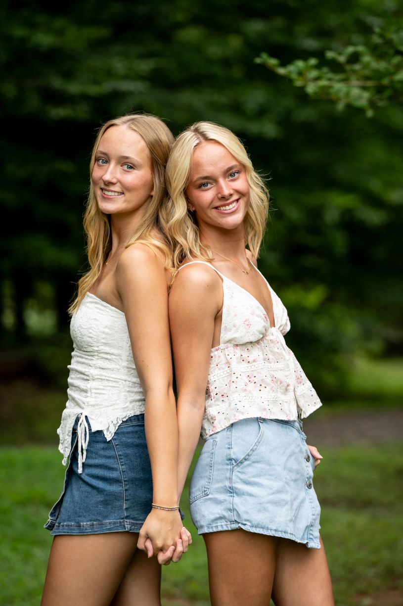 Two friends in summer outfits standing back-to-back and holding hands outdoors.