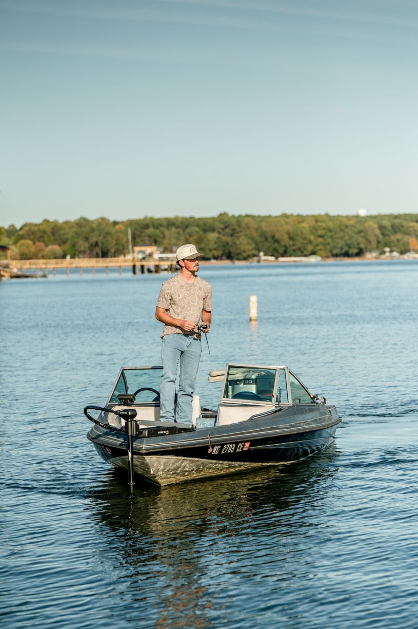 A small fishing boat floats with a senior boy on tranquil waters with trees visible along the shoreline.