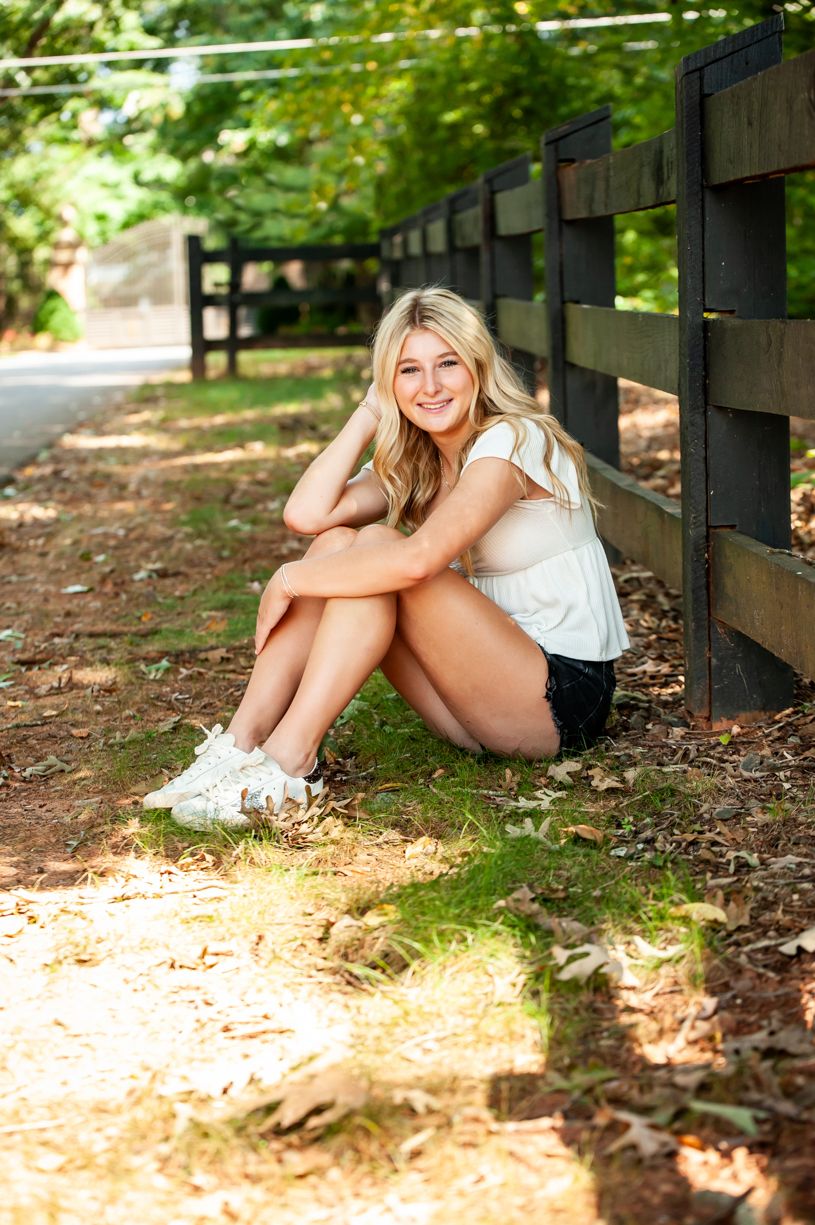 Senior girl sitting along rustic wooden fence in natural lighting.